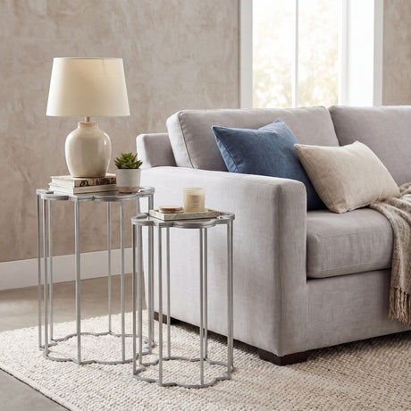 Hill Interiors Quatrefoil mirrored set of two nesting side tables in silver metal, styled beside a grey sofa with a lamp, books and candle on top.