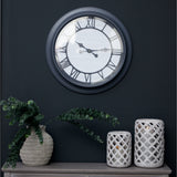 Hill Interiors 59cm round wall clock with silver frame and Roman numerals on a glass face, mounted on a dark wall above a console with vase and lanterns.