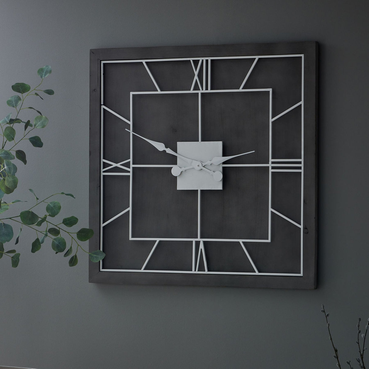 Hill Interiors Grey Square Metal Wall Clock with slim frame, open face, Roman numerals and slender hands, mounted on a dark grey wall beside foliage.