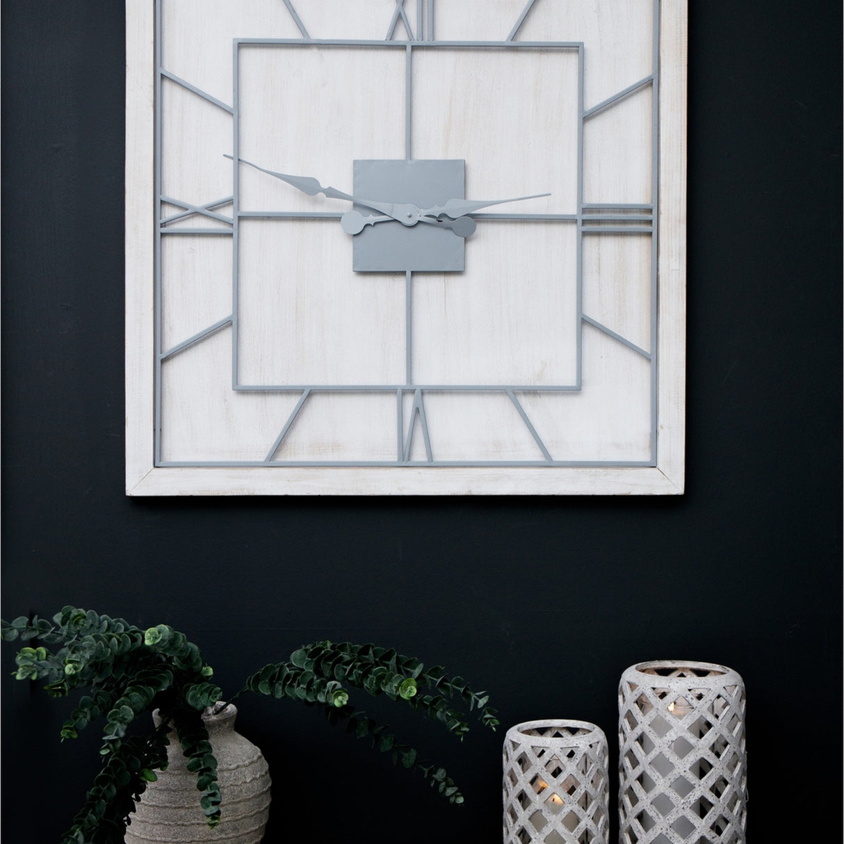 Hill Interiors White Square Large Wall Clock with slim metal frame and grey Roman numerals, mounted on a dark wall above eucalyptus and lanterns.