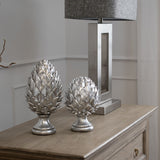 Two Hill Interiors silver ceramic pinecone finials with layered scaled forms on a light wood console beside a chrome table lamp with grey shade.