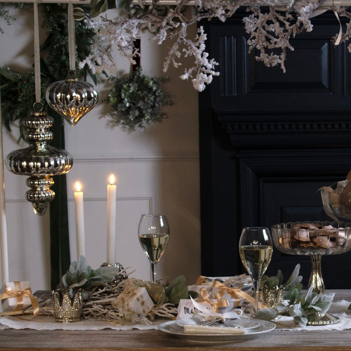 Antique silver glass baubles by Hill Interiors hanging from a garland above a festive table set with candles, wine glasses and gold-ribboned gifts.