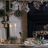Antique silver glass baubles by Hill Interiors hanging from a garland above a festive table set with candles, wine glasses and gold-ribboned gifts.