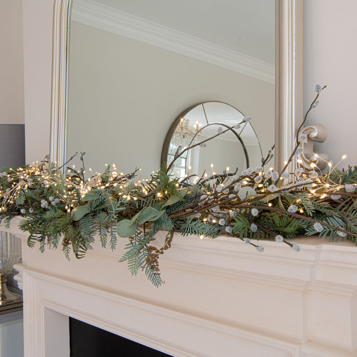 Hill Interiors warm white LED cluster string lights draped along a cream mantelpiece with foliage, pussy willow stems, and a framed mirror above.