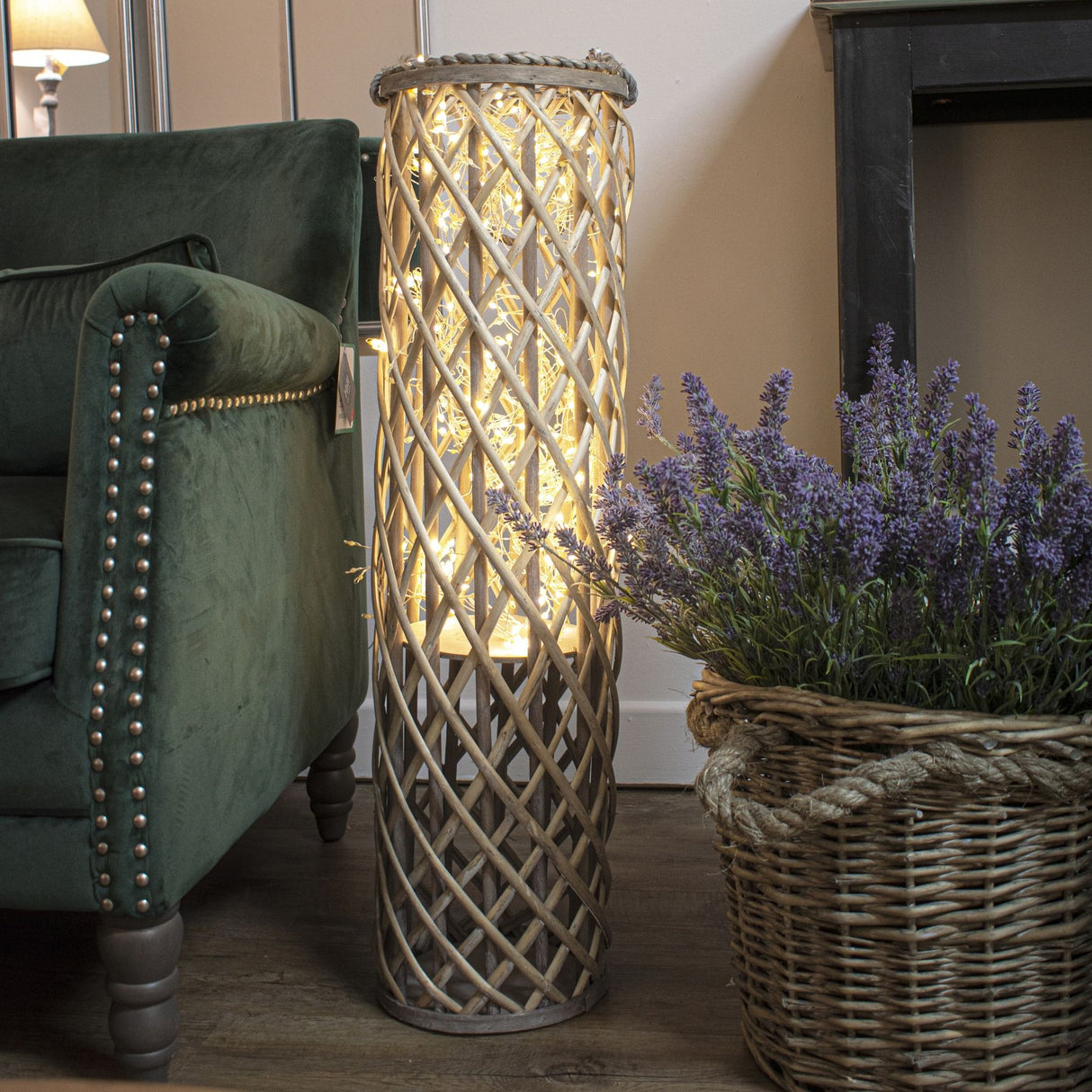 Hill Interiors LED cluster string lights glowing warm white inside a tall woven willow lantern beside a green velvet armchair and lavender basket.