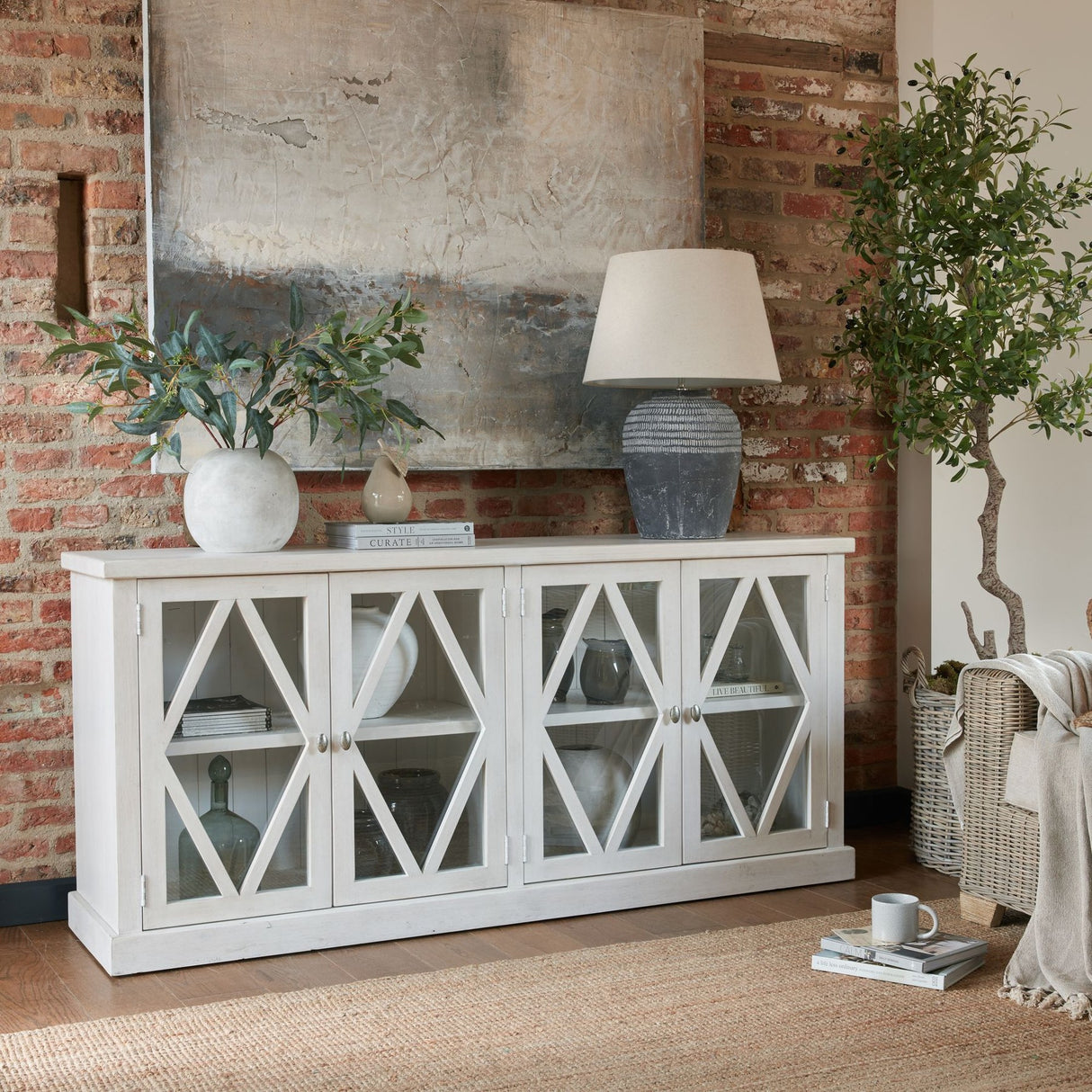 Hill Interiors Small Globe Ceramic Vase in beige with olive stems, styled on a white sideboard against an exposed brick wall with textured artwork.