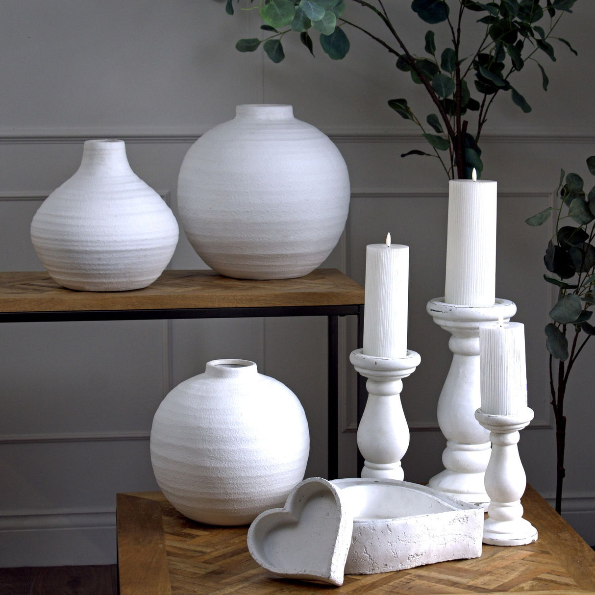 Hill Interiors matt white ceramic vase in a rounded spherical form, displayed on a wooden table with white candlesticks, larger vases and eucalyptus stems.