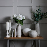 Hill Interiors matt white ceramic vase with rounded body beside candles, books, and tall stone vases with hydrangeas and eucalyptus on a wooden console.