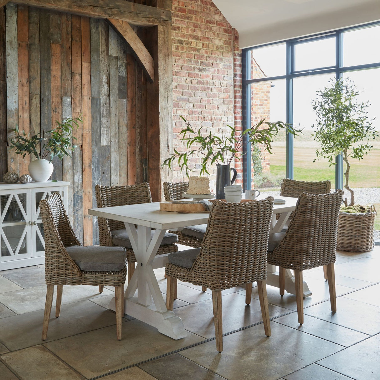 Hill Interiors matt white ceramic vase with foliage stems on a console beside a rustic wood-clad wall in a brick-walled dining room.