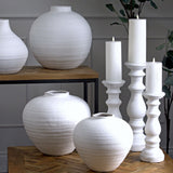 Hill Interiors Small Matt White Ceramic Column Candle Holder displayed with two taller matching holders, lit candles and textured white vases on wooden tables.