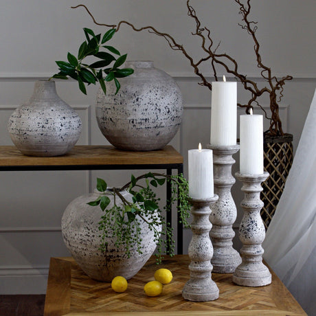 Hill Interiors Small Stone Ceramic Column Candle Holders in beige with lit white pillar candles, grouped with stone-effect vases and foliage on wood tables.