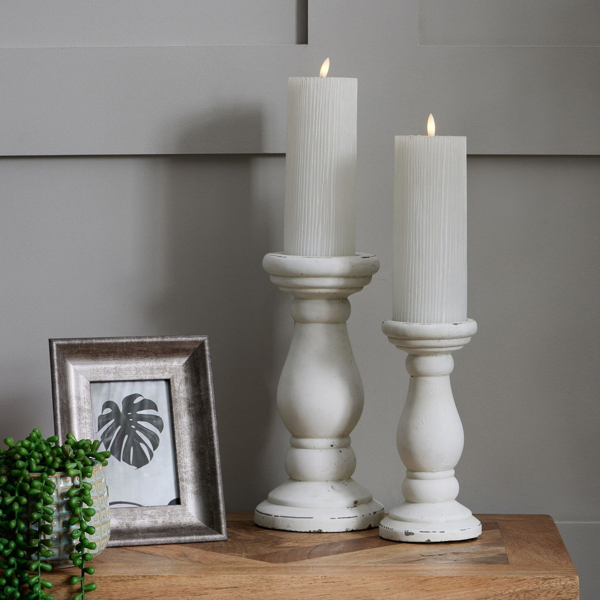 Hill Interiors matt white ceramic candle holders in two sizes with lit pillar candles, beside a silver photo frame and trailing plant.