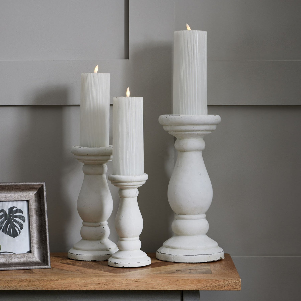 Hill Interiors matt white ceramic candle holders in three heights with lit pillar candles, arranged on a wooden console beside a framed print.