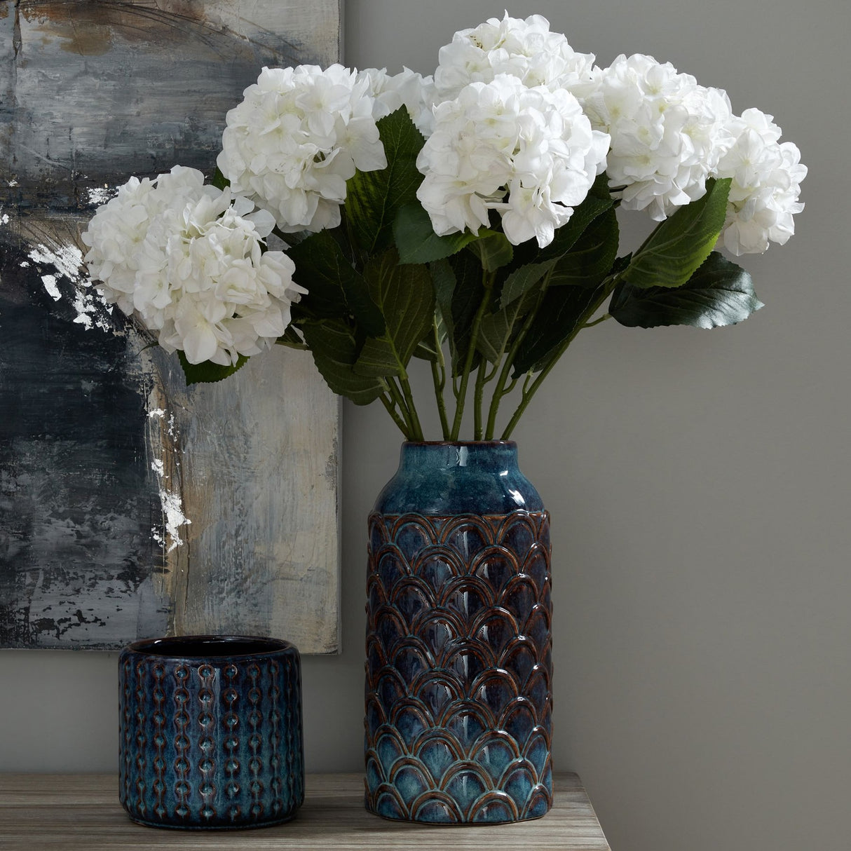 Hill Interiors Large Indigo Scalloped Ceramic Vase in blue holding white hydrangea stems, styled on a console beside a smaller ridged pot and abstract artwork.