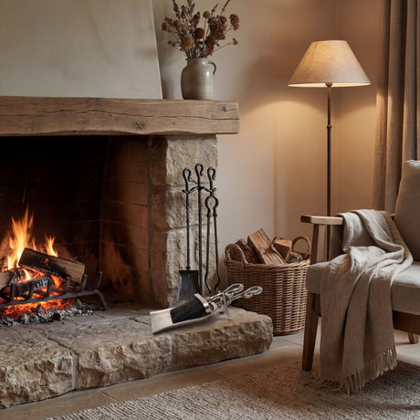 Hill Interiors Antique Pewter Crook Handled Hearth Tidy in silver with brush resting on a stone hearth beside a lit fireplace, log basket and floor lamp.