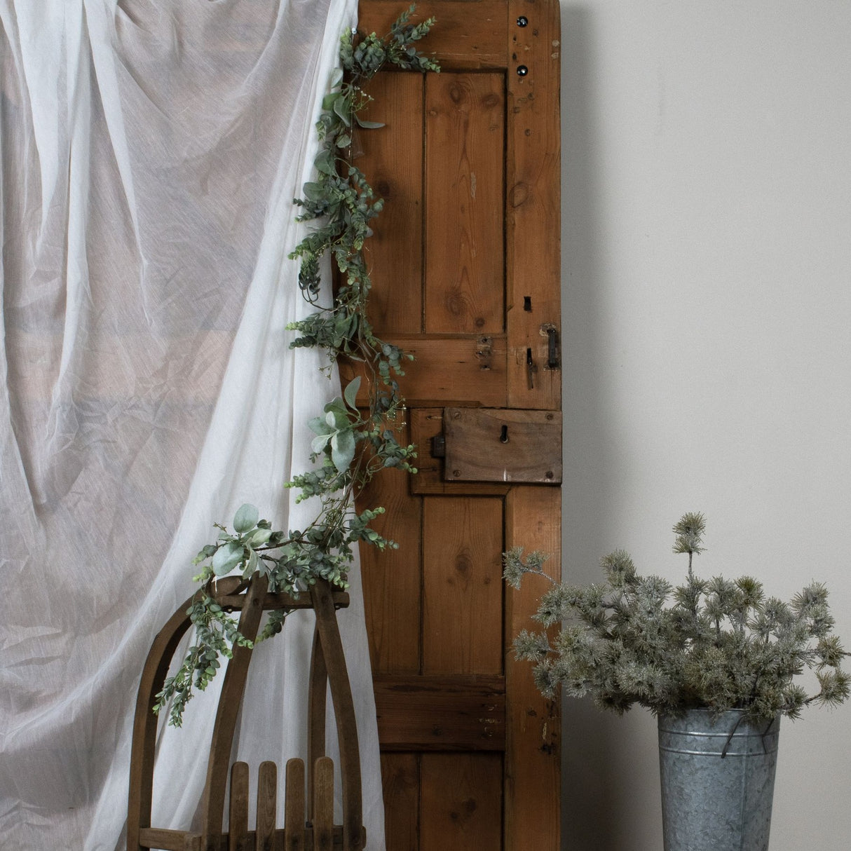 Hill Interiors LED winter garland with eucalyptus and lamb's ear draped over a rustic wooden door beside a sheer curtain and a zinc pot of foliage.