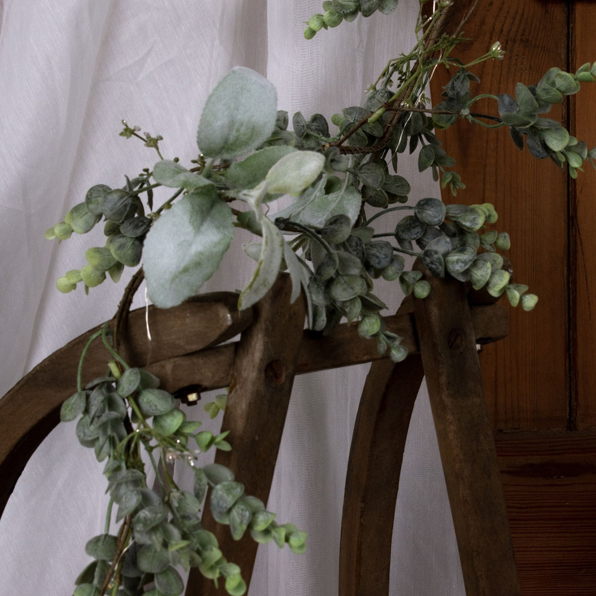 Hill Interiors LED winter garland with eucalyptus sprigs and lamb's ear leaves draped over a wooden chair against sheer white fabric.
