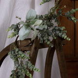Hill Interiors LED winter garland with eucalyptus sprigs and lamb's ear leaves draped over a wooden chair against sheer white fabric.