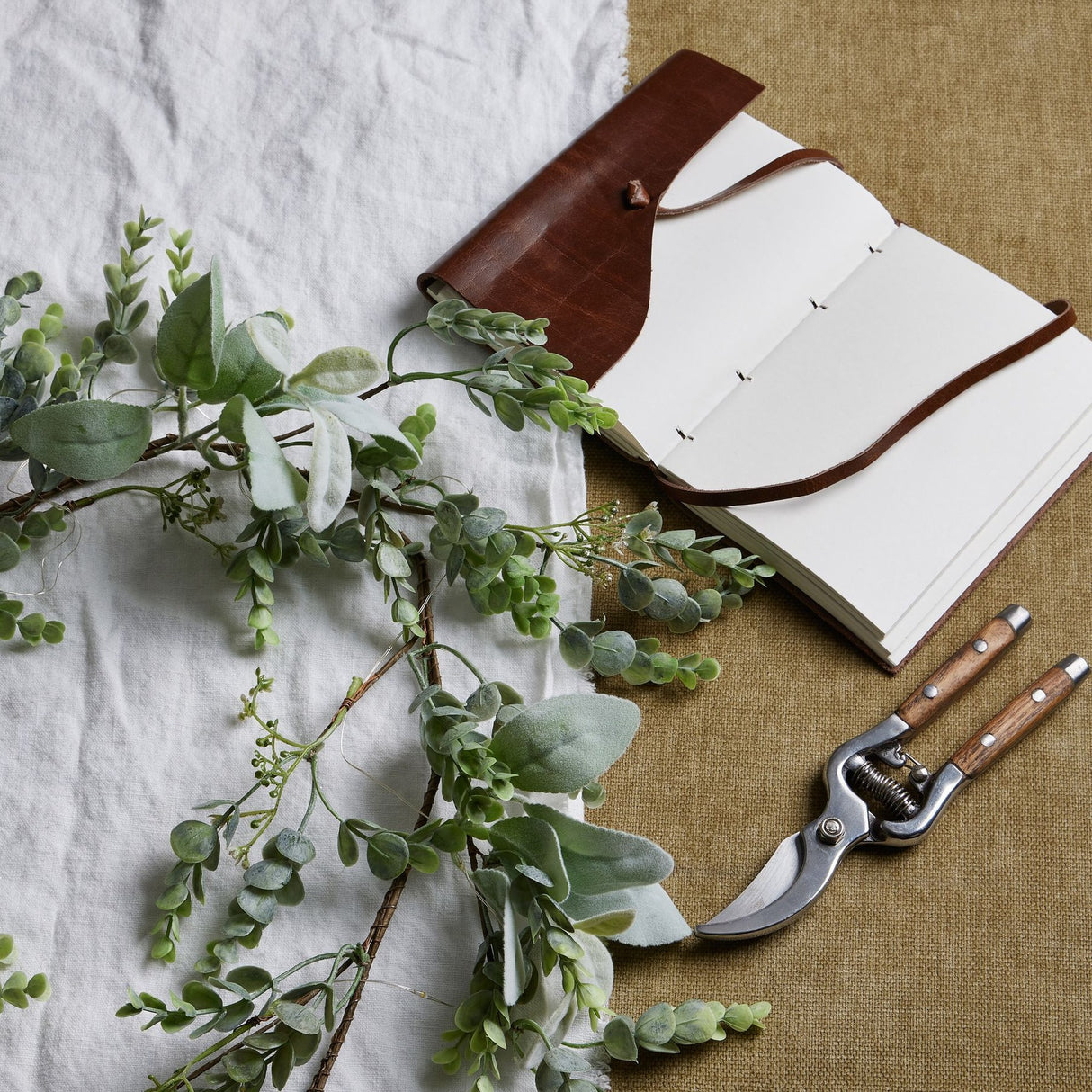 Hill Interiors LED winter garland with faux eucalyptus and lamb's ear styled beside an open leather-bound journal and wooden-handled secateurs on linen.