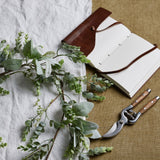 Hill Interiors LED winter garland with faux eucalyptus and lamb's ear styled beside an open leather-bound journal and wooden-handled secateurs on linen.