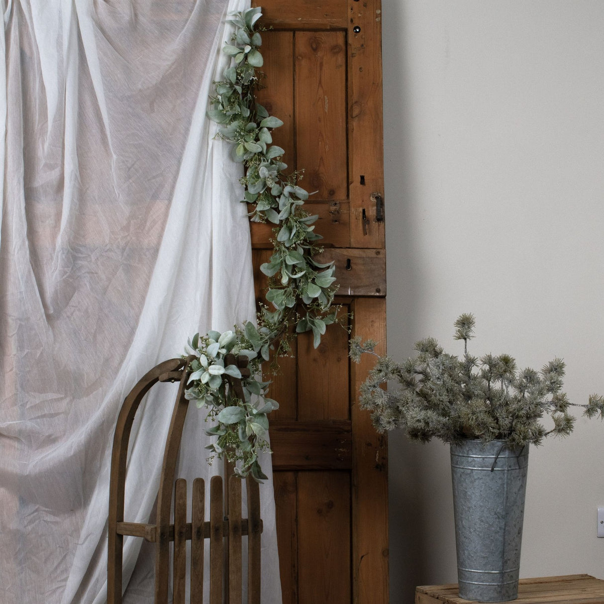 Hill Interiors Winter Garland with Lamb's Ear and Wax Flower draped along a rustic wooden door beside a linen curtain and a wooden chair.