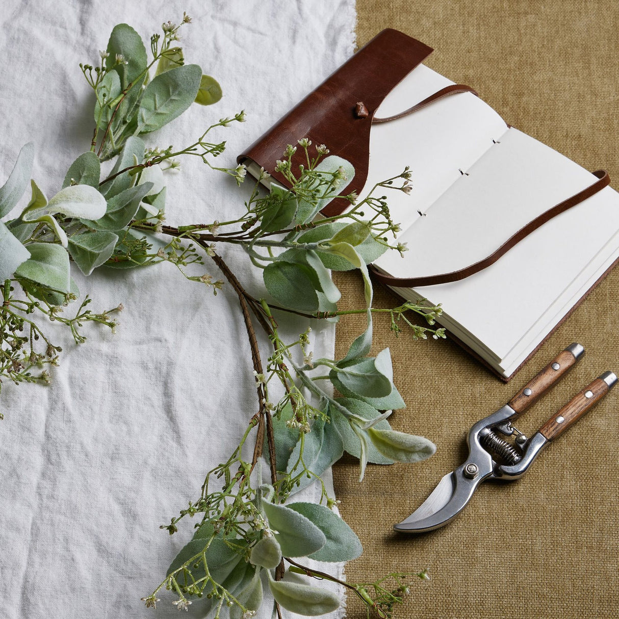 Hill Interiors Winter Garland with Lamb's Ear and Wax Flower in green, draped on linen beside a leather journal and wooden-handled secateurs.