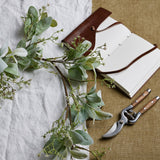 Hill Interiors Winter Garland with Lamb's Ear and Wax Flower in green, draped on linen beside a leather journal and wooden-handled secateurs.