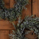 Close-up of Hill Interiors Frosted Pine Wreath with Pinecones in white, showing frosted branches, small pinecones and silvered berries against a wooden door.