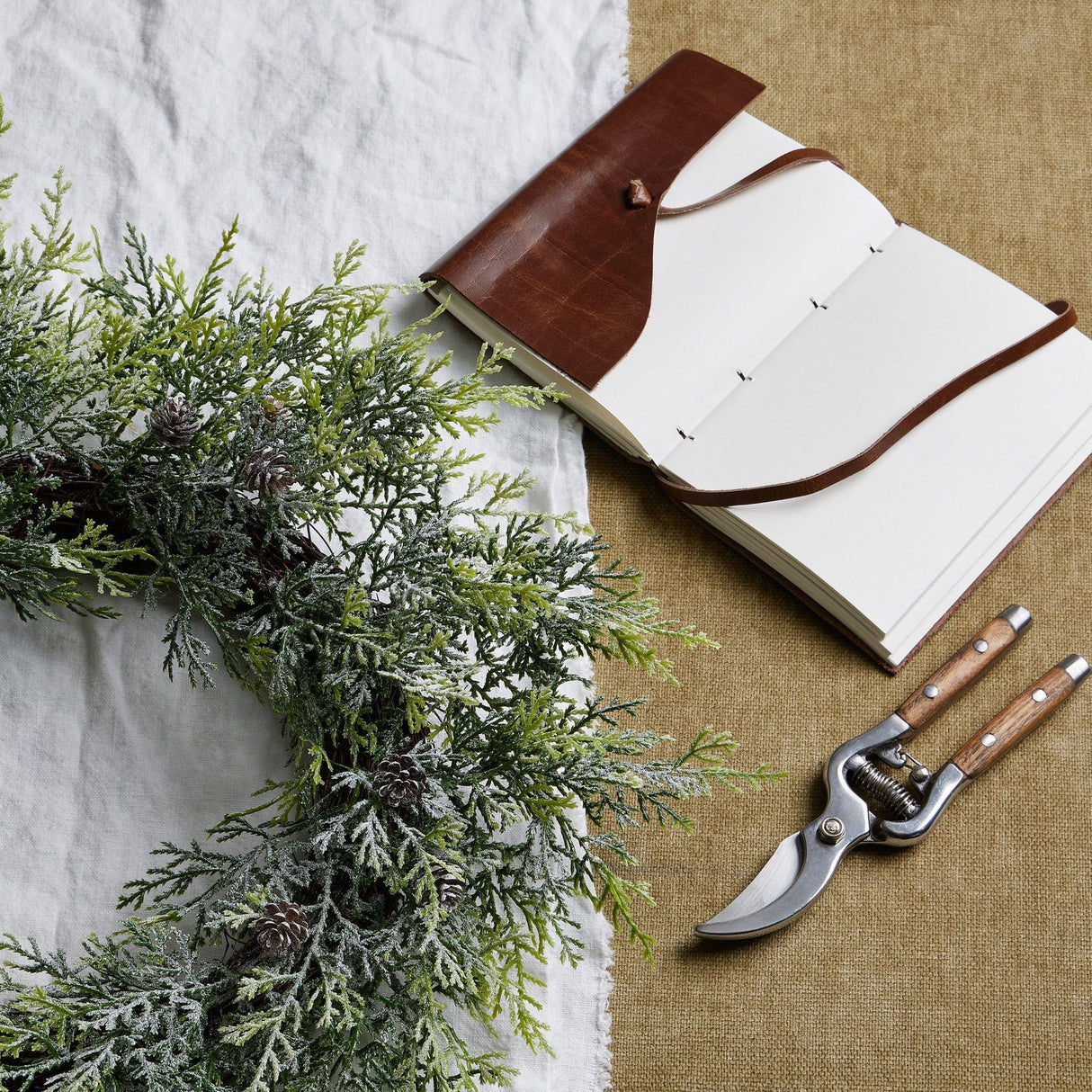Hill Interiors Frosted Pine Wreath with Pinecones on a white cloth beside an open leather-bound journal and wooden-handled secateurs on hessian.