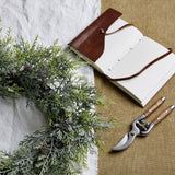 Hill Interiors Frosted Pine Wreath with Pinecones on a white cloth beside an open leather-bound journal and wooden-handled secateurs on hessian.