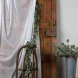 Hill Interiors Frosted Pine and Eucalyptus Garland in white draped over a wooden sled and along a rustic pine door beside a sheer curtain and pine bucket.