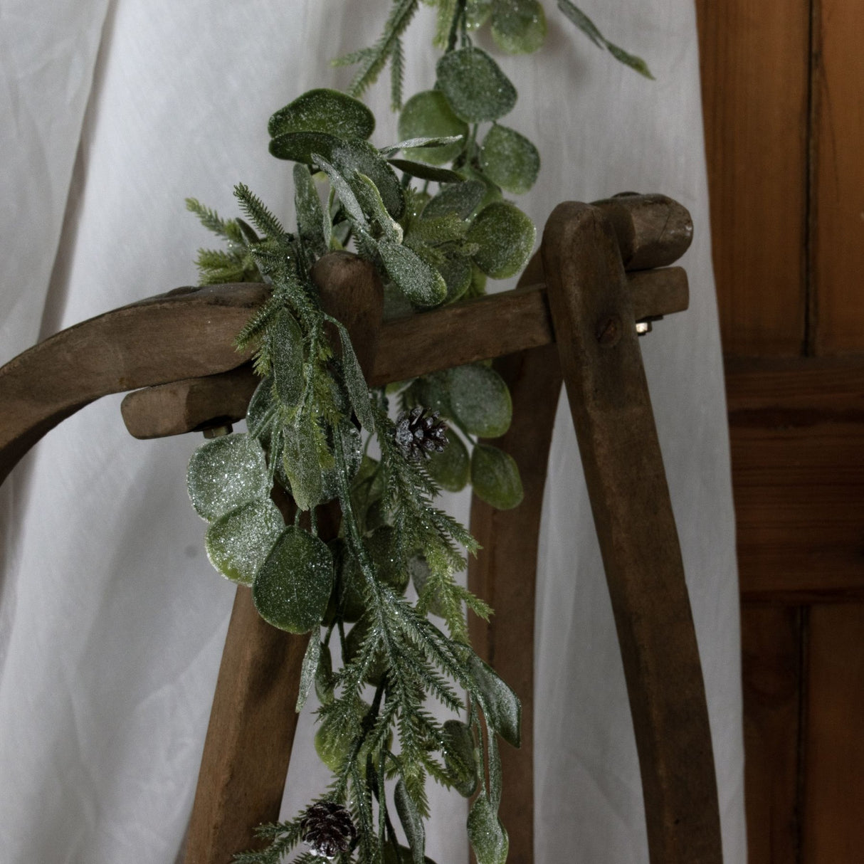 Hill Interiors Frosted Pine and Eucalyptus Garland in white draped over a rustic wooden chair, showing mixed foliage, pinecones and a light frosted finish.