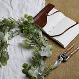 Hill Interiors Frosted Pine and Eucalyptus Garland in white draped on linen beside a leather journal and wooden-handled secateurs on hessian.