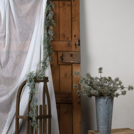 Hill Interiors Frosted Pine Garland draped over a wooden sled and rustic door beside a galvanised bucket of pine foliage in a neutral setting.