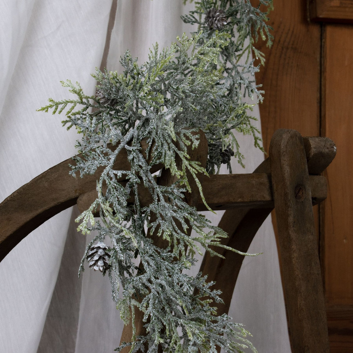Hill Interiors Frosted Pine Garland with Pinecones draped over a rustic wooden chair against a soft white fabric backdrop.