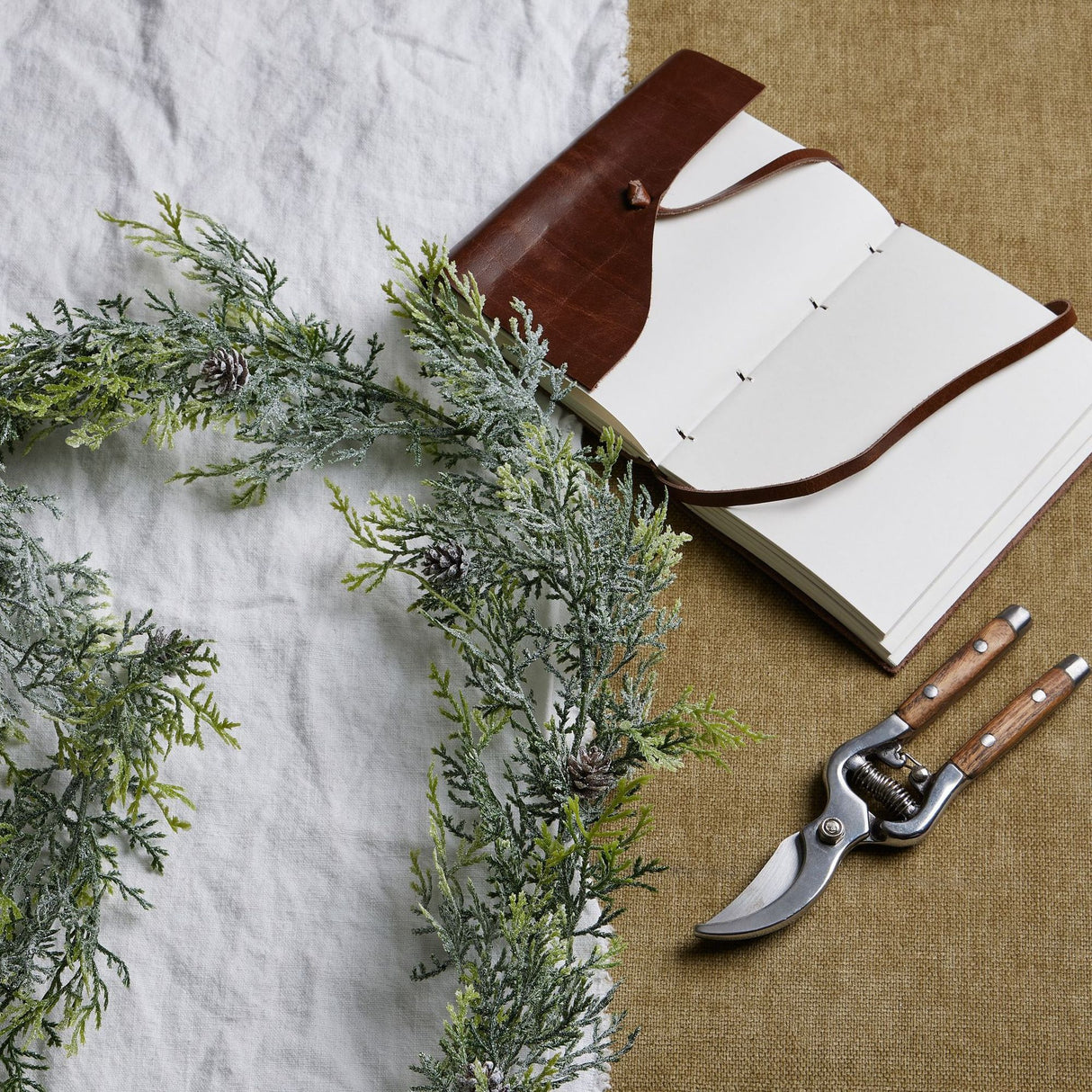 Hill Interiors Frosted Pine Garland draped on white linen beside an open leather-bound journal and wooden-handled secateurs on hessian.