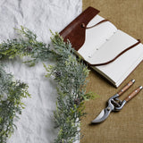 Hill Interiors Frosted Pine Garland draped on white linen beside an open leather-bound journal and wooden-handled secateurs on hessian.