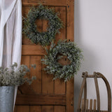 Hill Interiors Frosted Pine and Eucalyptus Wreath displayed with a second pine wreath on a rustic wooden door, styled beside a linen drape and wooden sledge.
