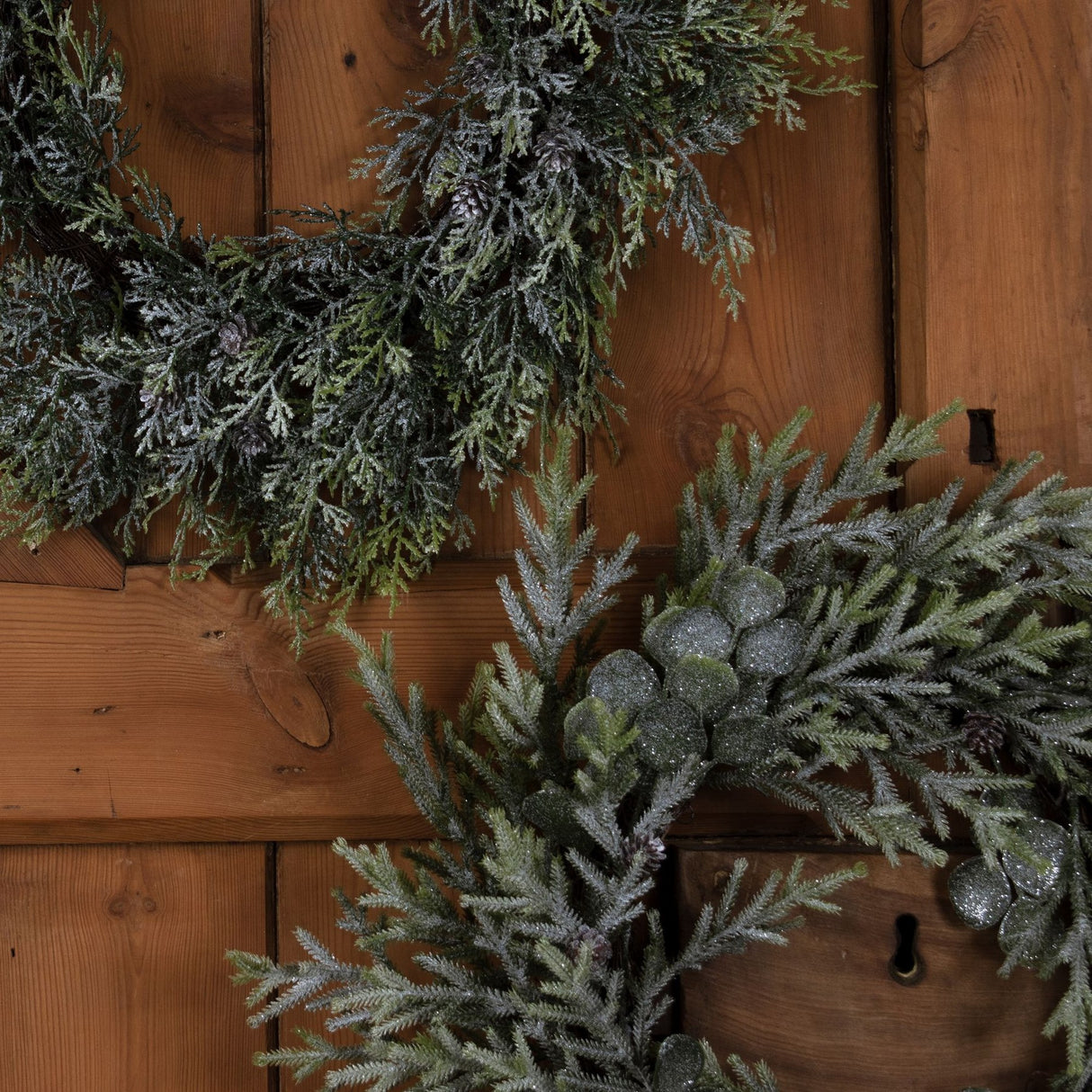 Hill Interiors Frosted Pine and Eucalyptus Wreath detail showing frosted green foliage, pinecones and silvered berries against a wooden panelled door.