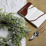 Hill Interiors frosted pine and eucalyptus wreath laid on white linen beside an open leather-bound journal and wooden-handled secateurs on hessian.