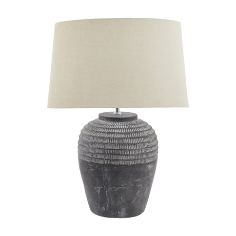 Hill Interiors Ferranock table lamp with carved stone-effect ceramic base in dark grey and tapered beige linen shade, shown on a plain white background.