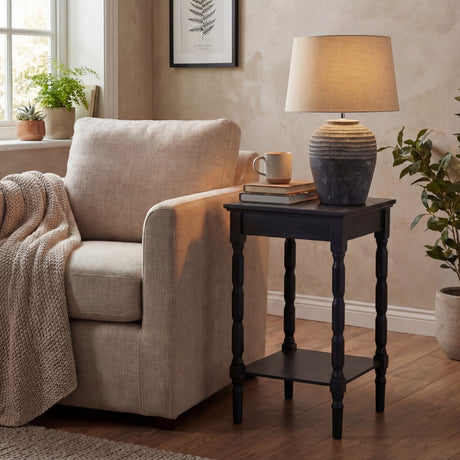 Hill Interiors Ferranock carved stone-effect ceramic table lamp in beige with tapered shade, styled on a black side table beside a linen armchair.