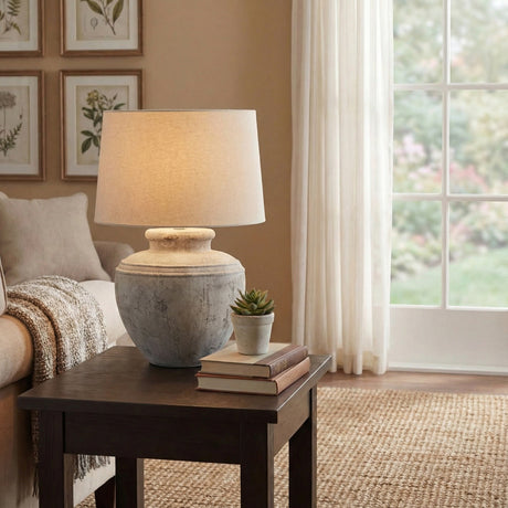 Hill Interiors Merenza ceramic table lamp with a rustic stone-toned urn base and cream drum shade, styled on a dark side table beside books and a succulent.