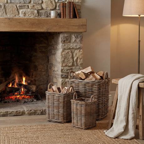 Hill Interiors Set of 3 Square Rattan Storage Baskets in brown holding firewood and kindling beside a rustic stone fireplace in a living room.