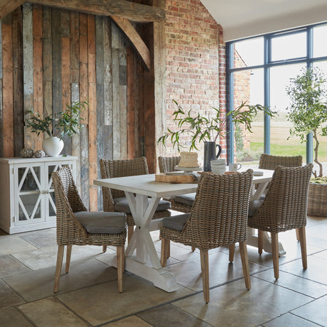 Hill Interiors Rattan Garden Dining Chairs in beige around a white trestle table in a rustic barn dining room with brick and timber walls.