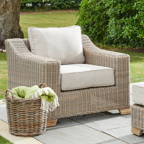 Hill Interiors Metal Outdoor Armchair in beige with woven frame and cream cushions, styled on a garden patio with a wicker basket and throw.