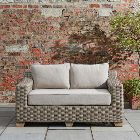Hill Interiors Rattan Two-Seater Garden Sofa in beige with woven frame and plush cushions, set on stone paving against a rustic brick wall.