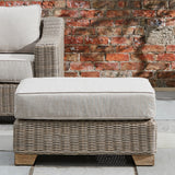 Hill Interiors Outdoor Foot Stool in beige with woven rattan-style base, wooden feet and a cushioned top, set on a patio against a brick wall.