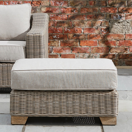Hill Interiors Outdoor Foot Stool in beige with woven rattan-style base, wooden feet and a cushioned top, set on a patio against a brick wall.
