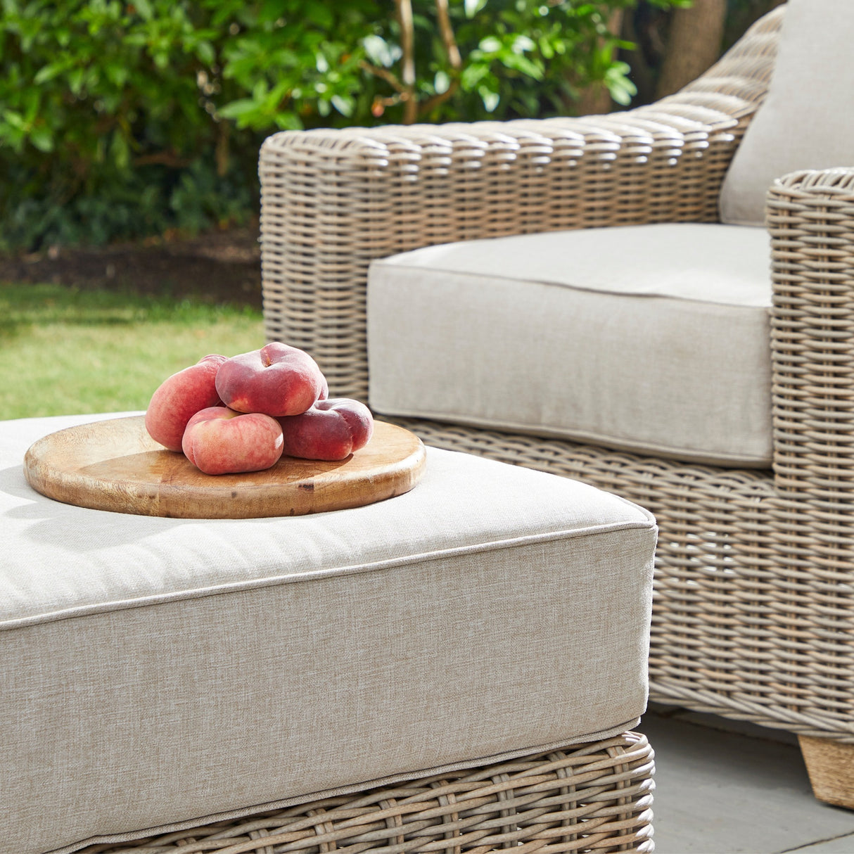 Hill Interiors beige outdoor foot stool with woven rattan base and cushioned top, styled with a wooden board of peaches beside a matching garden armchair.